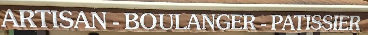 Gros plan sur la banne de la boutique : lettres blanches sur toile brune formant Artisan boulanger pâtissier.
